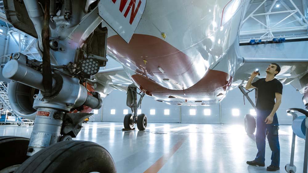In a Hangar Aircraft Maintenance Engineer/ Technician/ Mechanic Visually Inspects Airplane's Chassis and Body/Fuselage walking Under It.