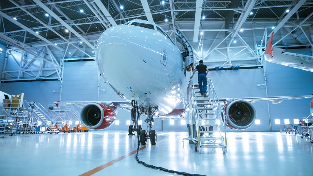 Brand New Airplane Standing in a Aircraft Maintenance Hangar while Aircraft Maintenance Engineer/ Technician/ Mechanic goes inside Cabin via Ladder/ Ramp.